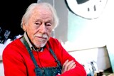The artist Guy Warren, a man in his late 90s with a apron on and arms crossed, sitting in his studio