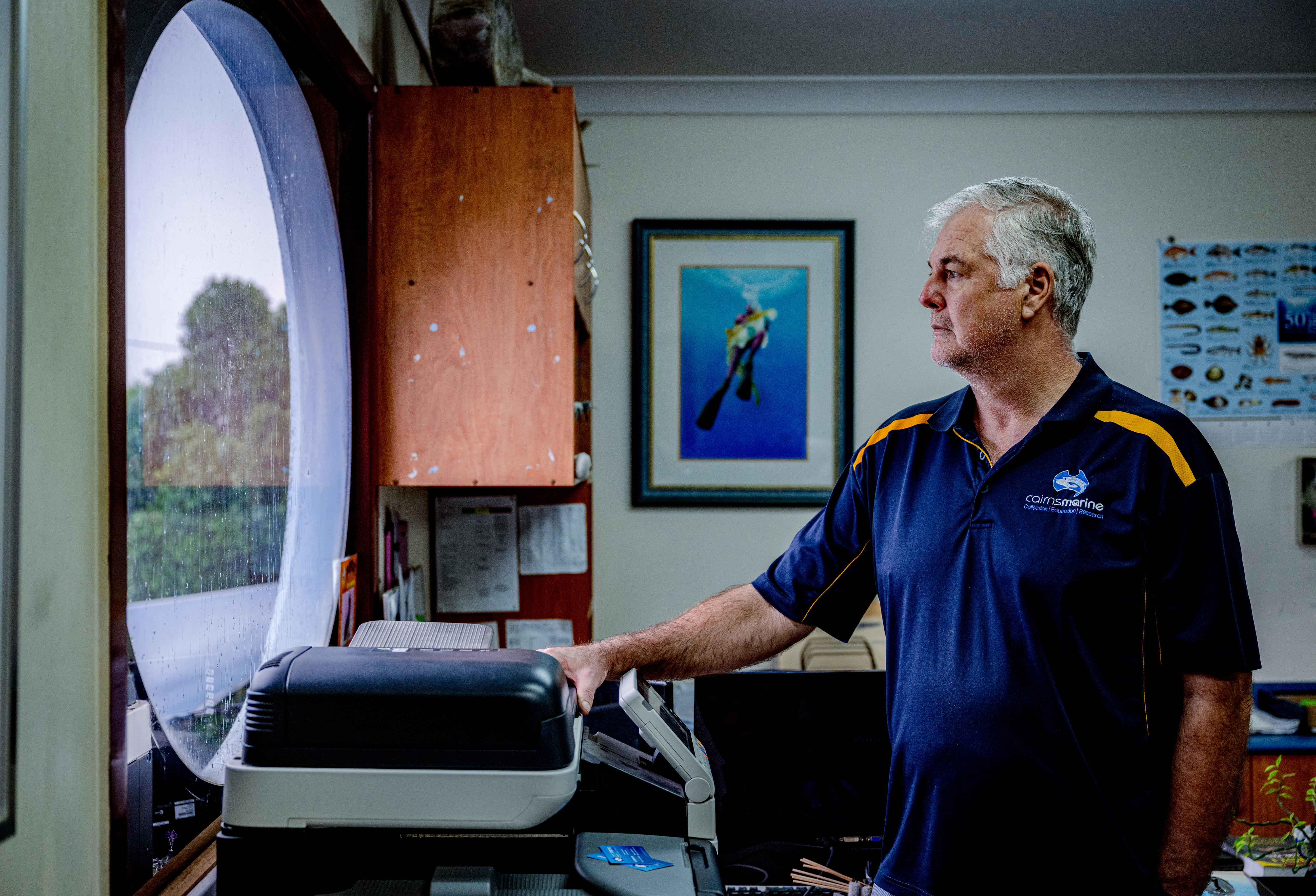 Lyle mira por una ventana redonda en su oficina. En la pared hay una fotografía de un buceador y un mapa de especies de peces.