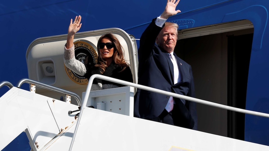 A tight shot man and a woman wave as they disembark from a plane and step onto the stairs.