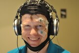 A smiling man wears a black, balaclava-style head covering with four electrodes visible and a earphone headset