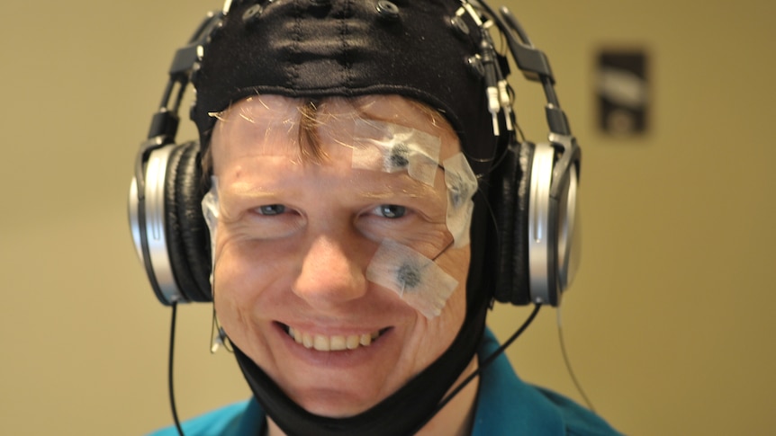 A smiling man wears a black, balaclava-style head covering with four electrodes visible and a earphone headset