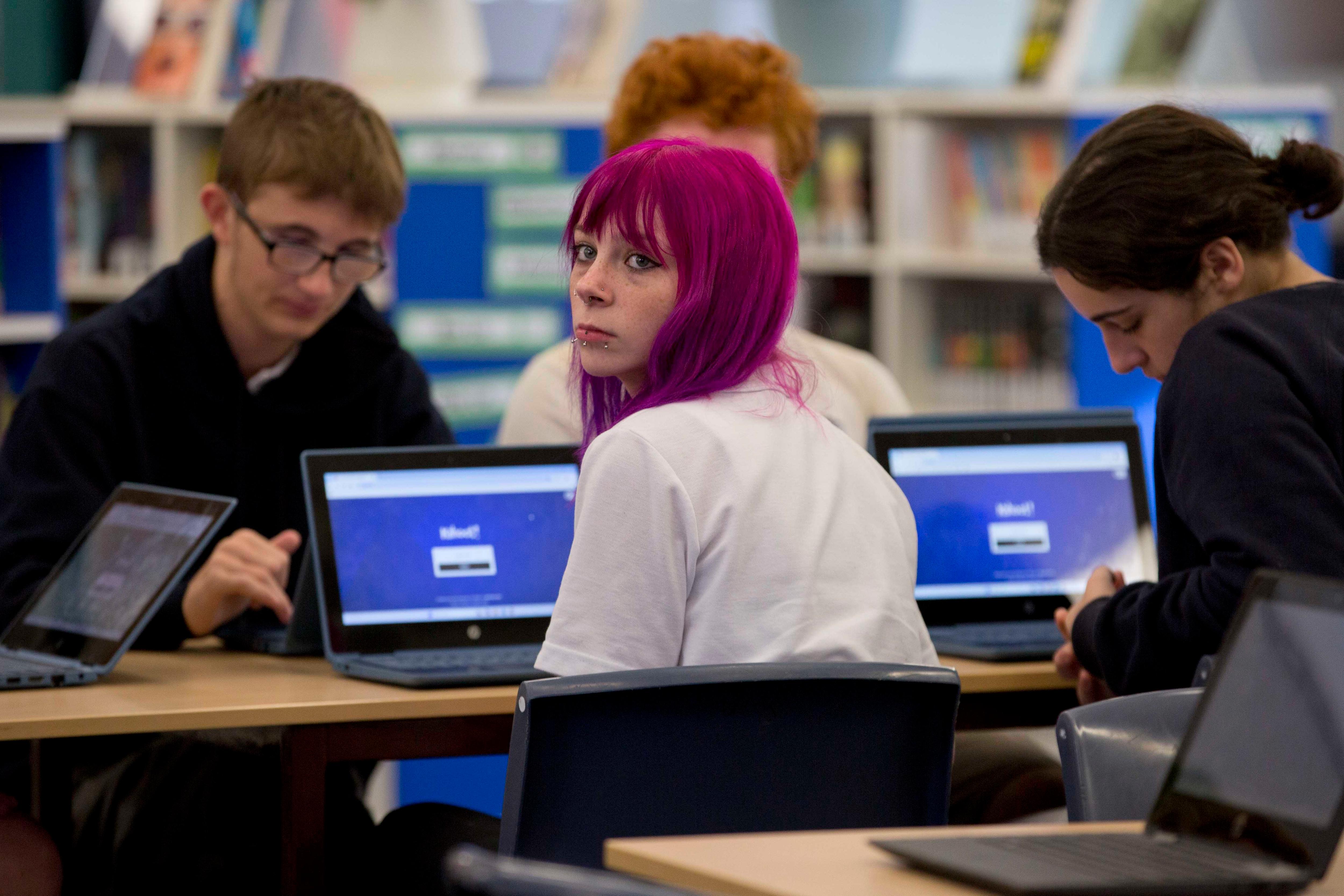 Students are sitting with their laptops in a library, focused on their screens in front of them.