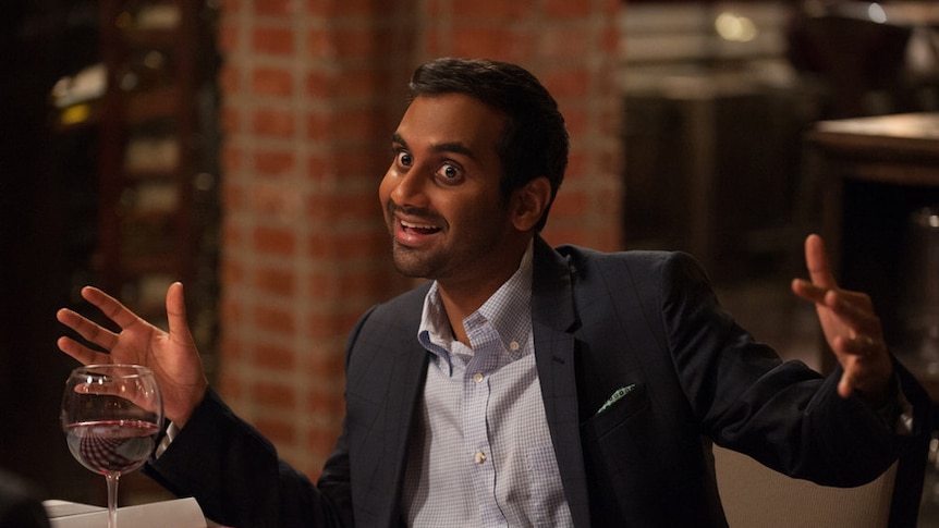 Aziz Ansari sits on a table drinking wine.