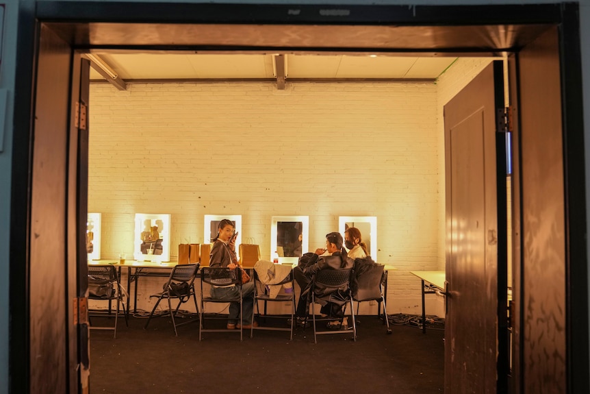 Three models sit backstage in a vast brick room lit with yellow light.