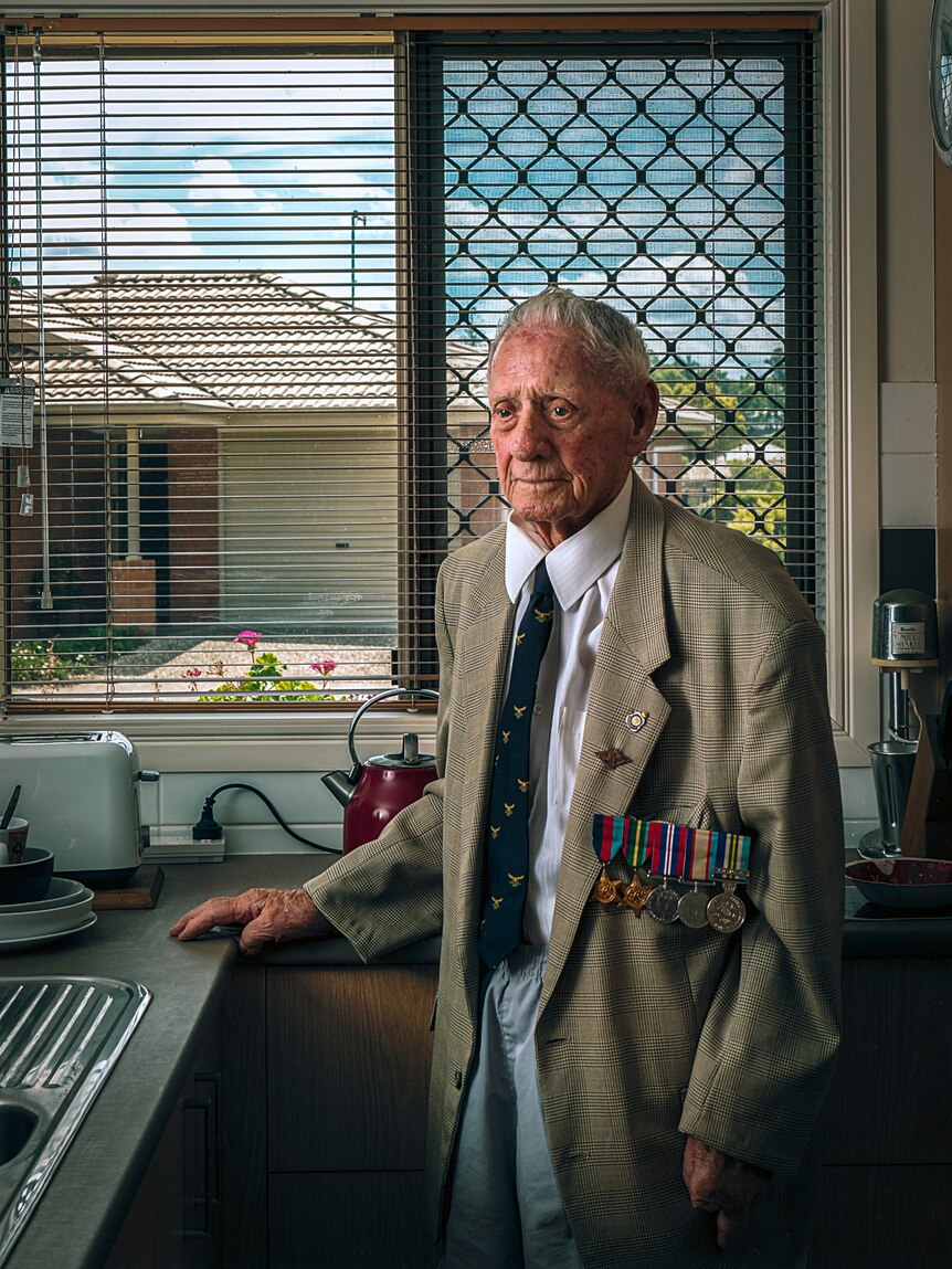 A photograph of an elderly man in his kitchen wearing a suit jacket, tie and military service medals.
