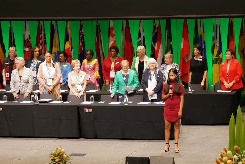 A group of women stands in front of flags of member nations in a conference hall.