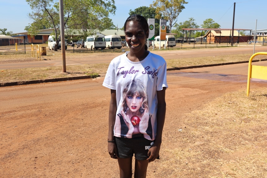 A woman in a Taylor Swift T-shirt smiles at the camera, Wadeye town in background