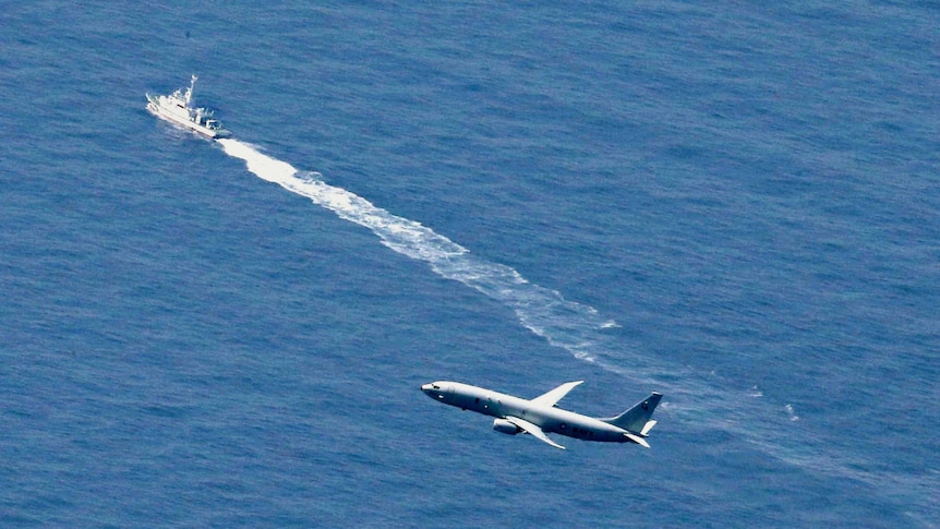 An aerial photo over the sea shows a military reconnaissance flying above a patrol boat on the water line in clear blue waters.