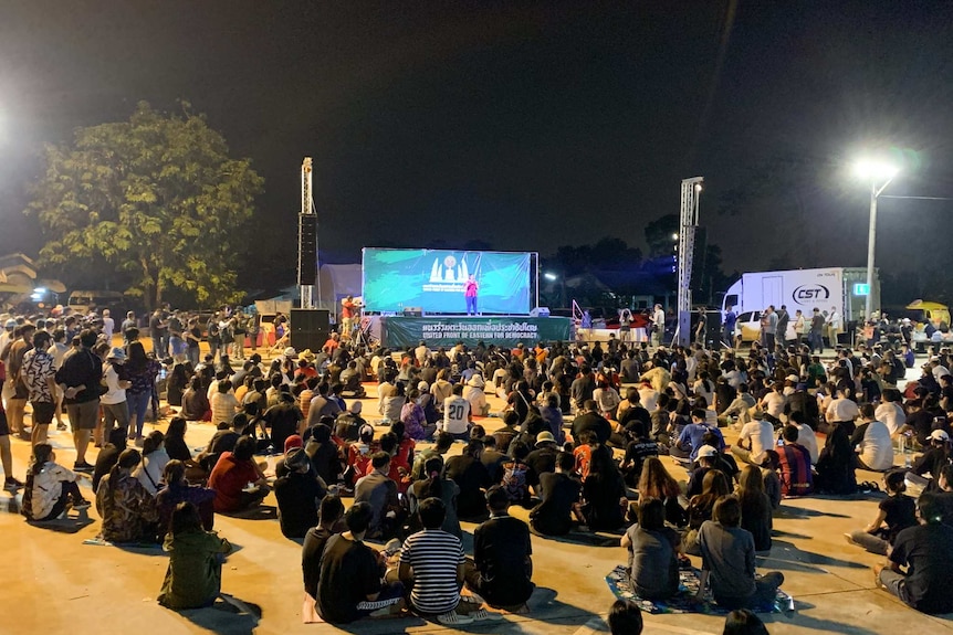 A crowd of people sit on the ground looking at a stage where a woman speaks.