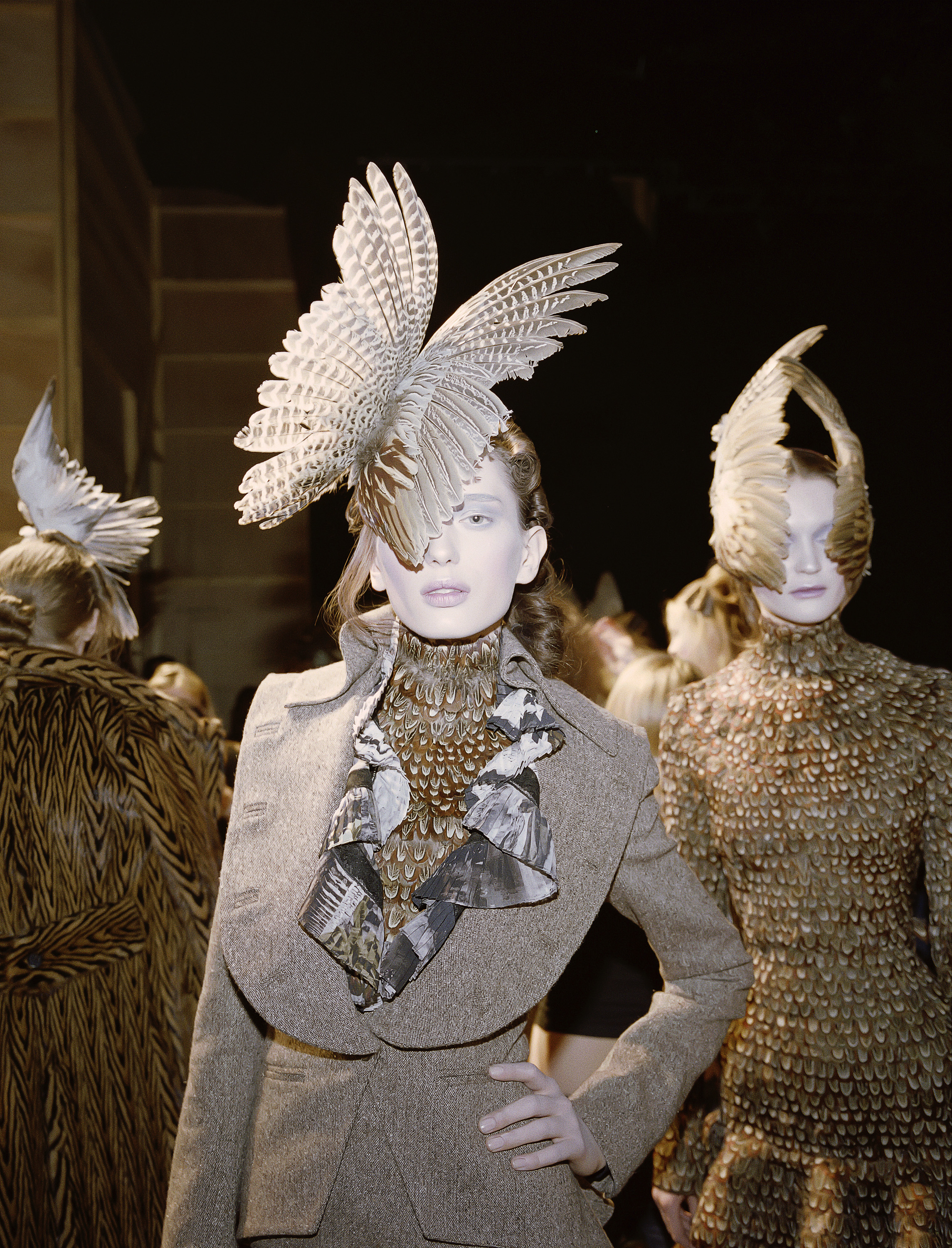 A model poses with one hand on her hip, wearing a feather headdress and unique blazer, her face powdered white