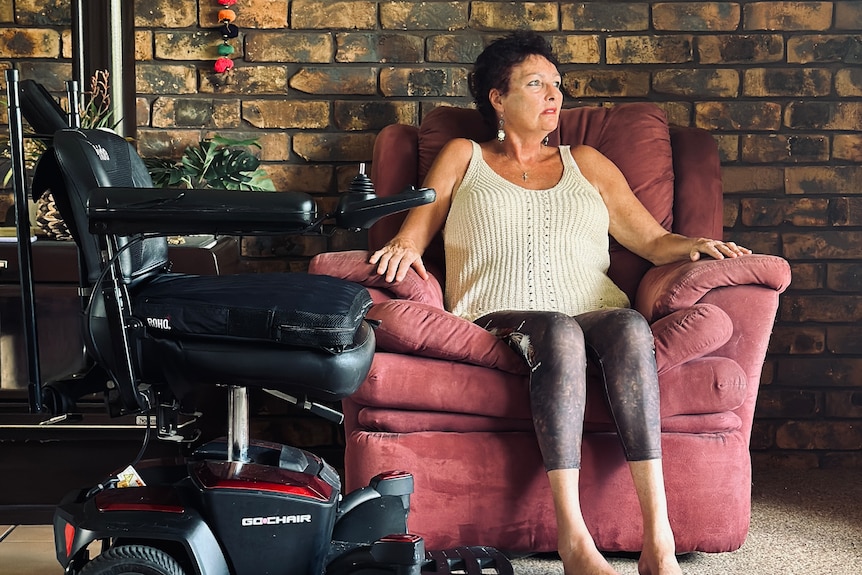 A woman sits on a couch inside a home. Next to her is her motorised wheelchair.