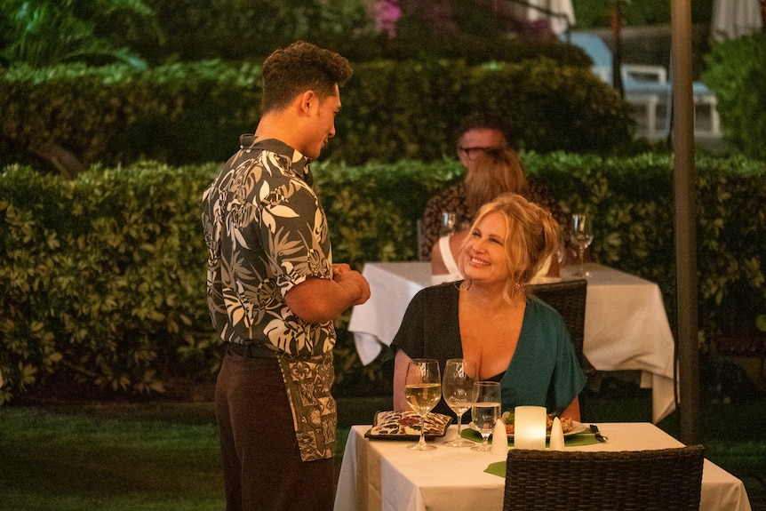 A young Native Hawaiian man takes the order of an older white woman with blonde hair sitting at a table