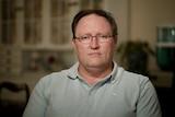 A man in a light blue polo and glasses looks at the camera looking despondent.