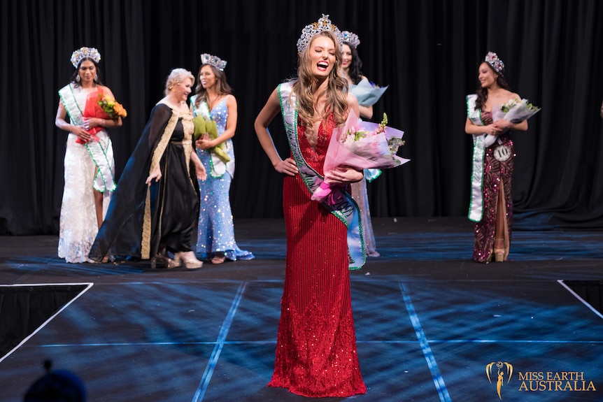 Sheridan Mortlock, wearing long red dress and silver crown, stands on a stage smiling widely, holding flowers.