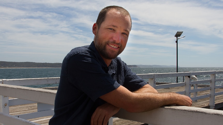 A man with short dark hair and a beard, wearing a navy shirt, standing at a jetty by the ocean.