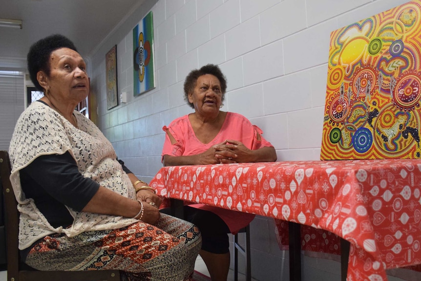 Two women sit at a table and speak to someone off camera. An Indigenous artwork sites on the table