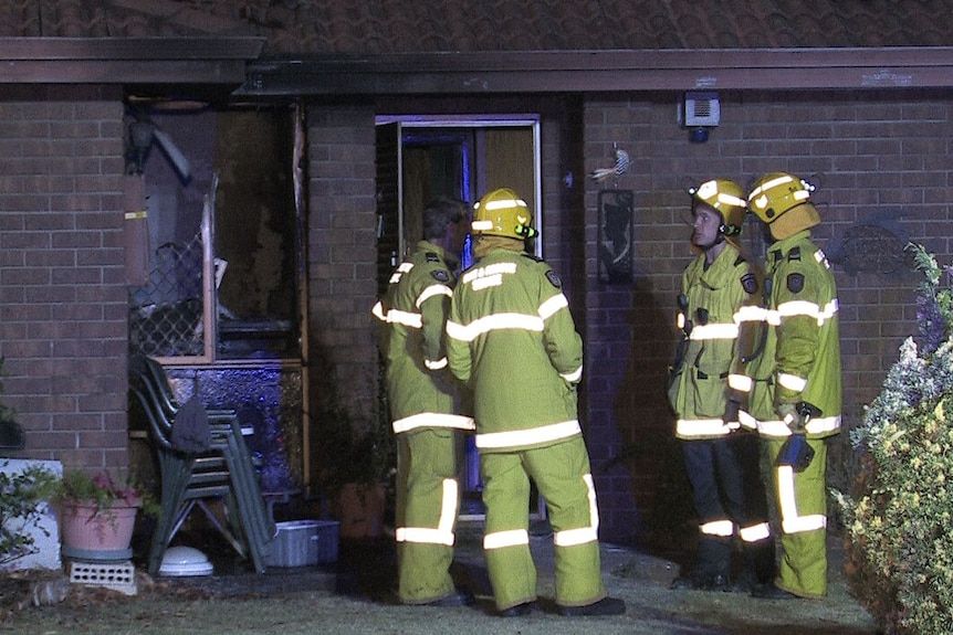Four firefighters stand outside the front door of a fire-damaged house, with a broken window visible.