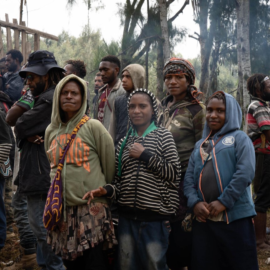 A group of mostly young PNG men and women stand in a village clearing in a forest, looking at the camera.