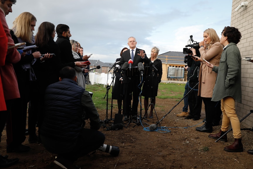 Morrison speaks at a microphone backed two women, with a stretch of mud between to banks of journalists