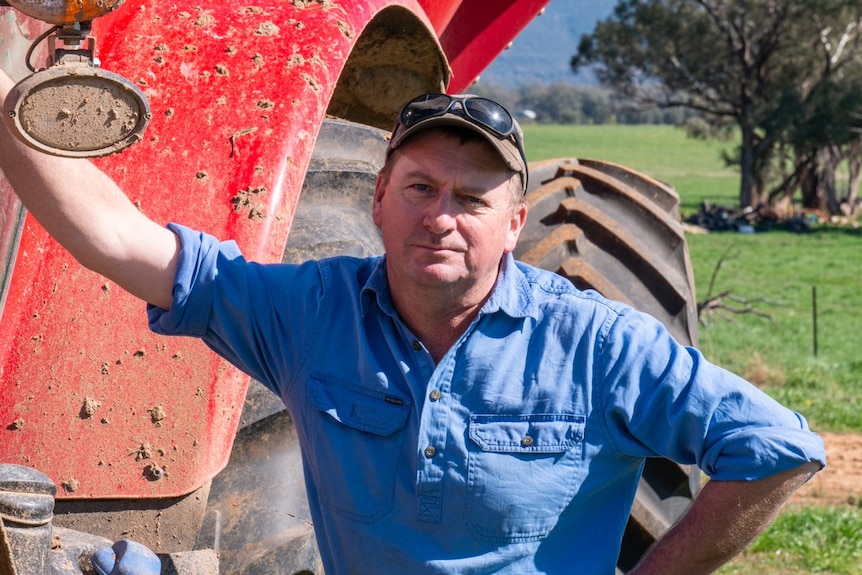Farmer, Chris Groves, standing in front of his dusty tractor on his farm in a paddock with a tree behind, looks serious.