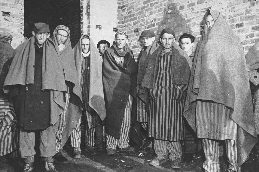 Black and white photo of men huddled together wrapped in blankets, wearing striped uniforms, looking gaunt and serious.
