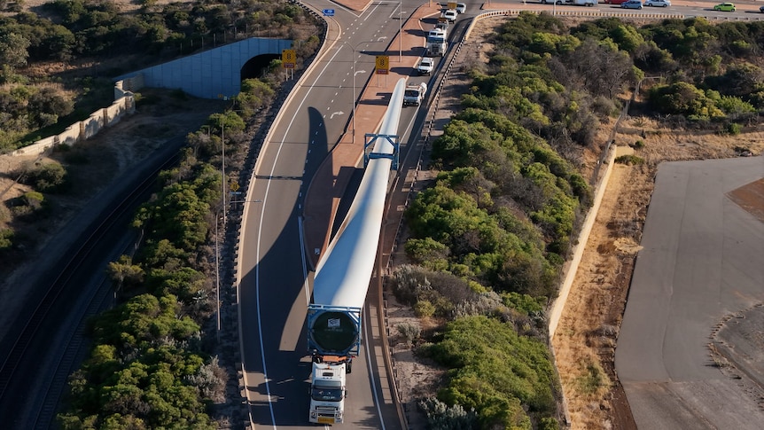 An aerial view of a truck carrying a giant wind turbine blade under escort.  
