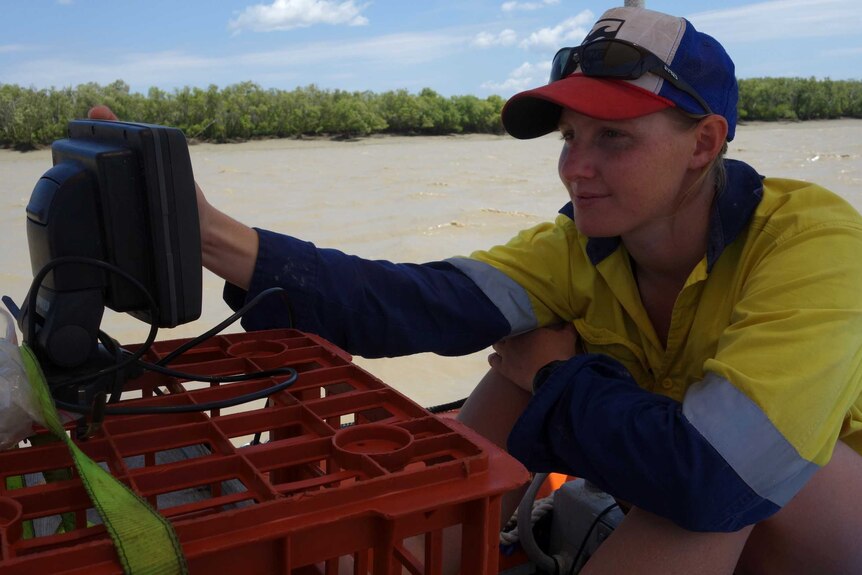 A young woman wearing a cap and high-vis gear looks at a small monitor that is perched on a milk crate.