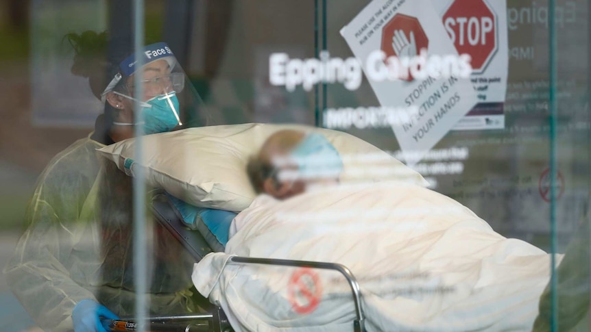 A man wearing a face mask in a hospital bed is wheeled out by a health worker in a face shield and mask.