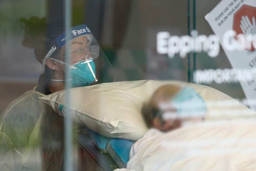 A man wearing a face mask in a hospital bed is wheeled out by a health worker in a face shield and mask.