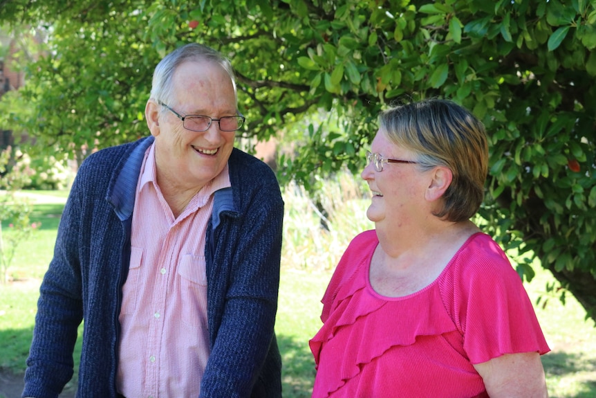 A elderly man wearing a cardigan smiles as he looks at an elderly woman who is wearing a pink shirt