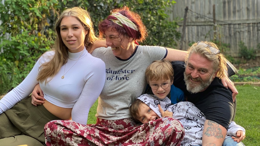 A mother holds her daughter, left, and two sons, middle and right, with her partner on the far right, in a garden.