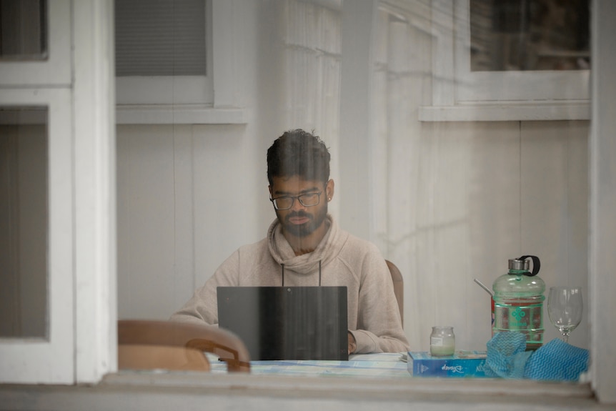 Arpit Charwal is seen through a window working at a laptop computer.