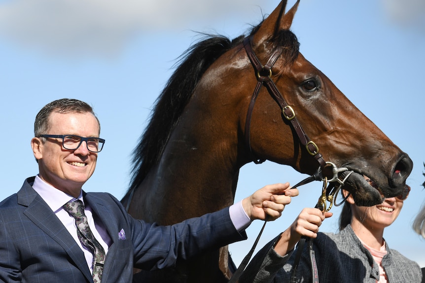 a man in a suit and glasses smiling and posing with a horse outdoors