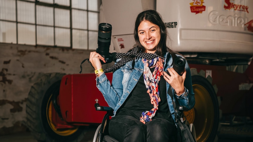 A woman in a wheelchair poses smiling with a long lens camera around her neck.