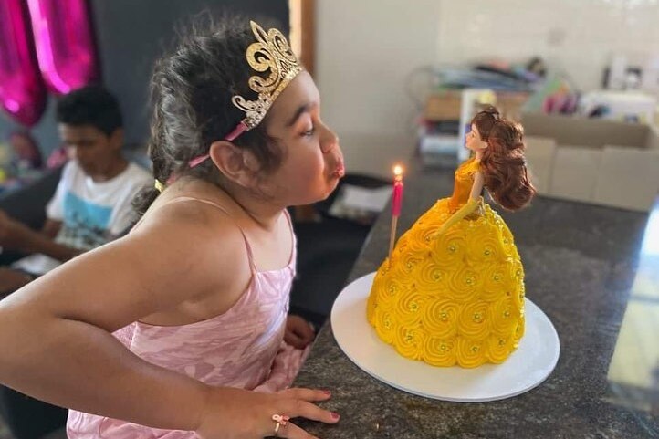 A girl in a pink dress and tiara blows out a single pink candle on a princess cake.