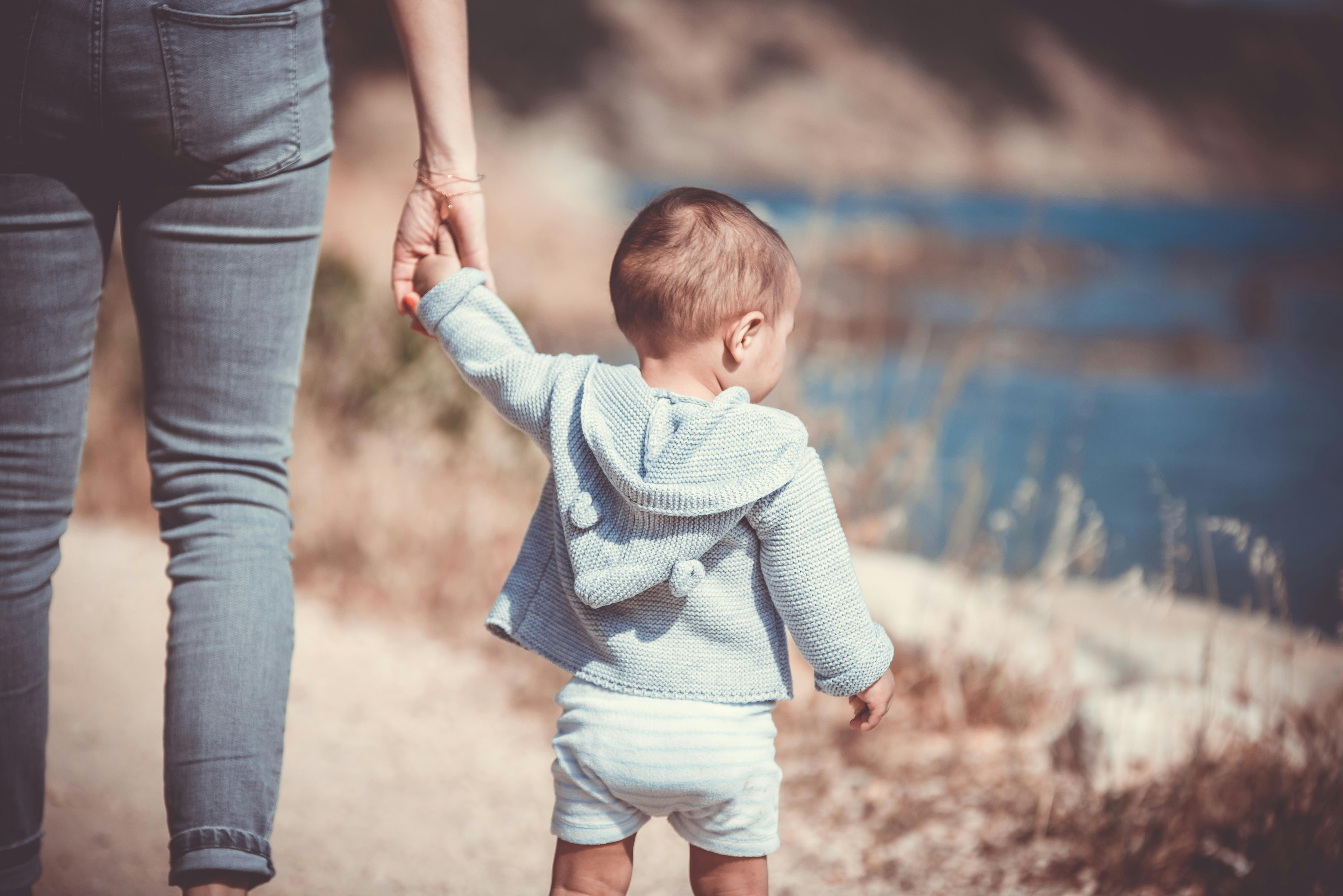 A mother holds her baby by the hand as they go for a walk along a pth by the water