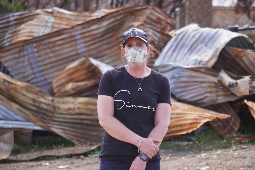 Jessica wears a black t-shirt and white mask, standing in front of burnt and destroyed corrugated iron. 