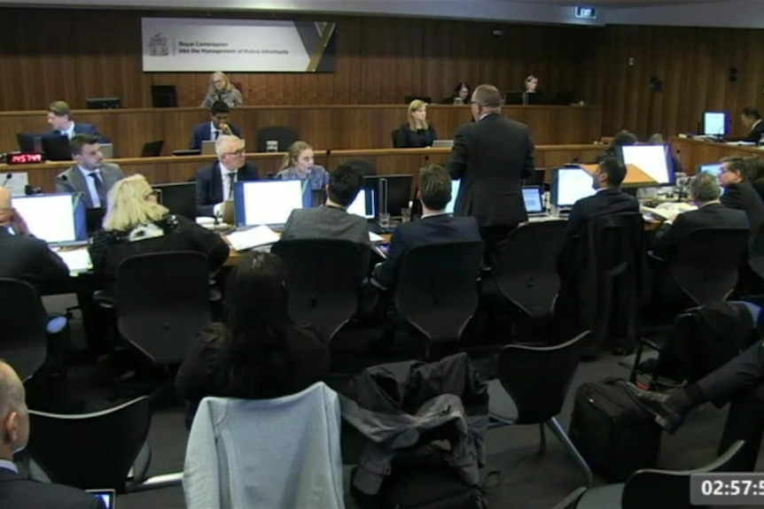 A screenshot of a web stream that shows lawyers sitting in a packed room, speaking to a commissioner.