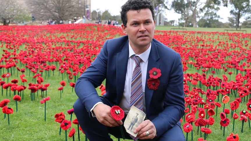 Phillip Johnson squats in a field of poppies, holding a poppy and an old photograph.