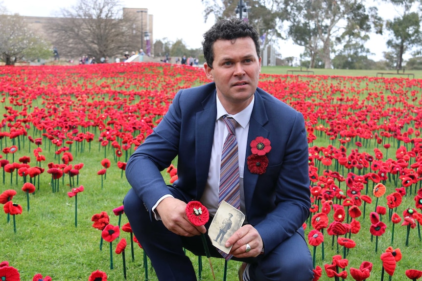 Phillip Johnson squats in a field of poppies, holding a poppy and an old photograph.