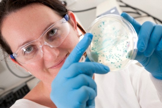 Woman in white lab coat and blue gloves holding up petri dish. 