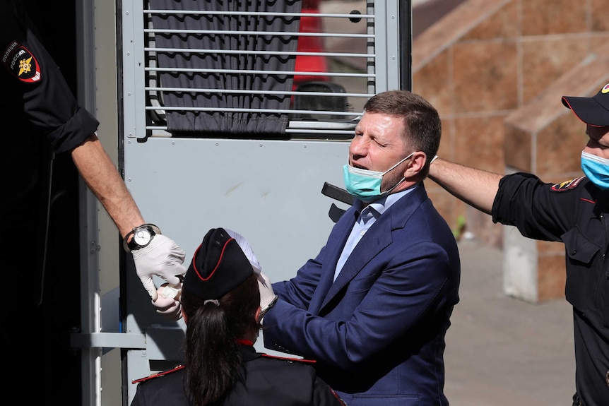 A handcuffed man in a navy suit squints as he is moved into the rear of a secure vehicle by two uniformed police officers.