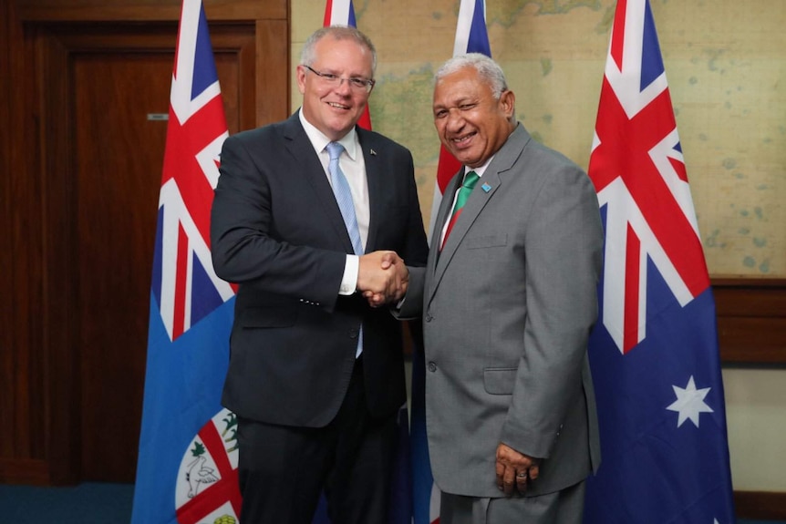 Scott Morrison and Frank Bainimarama shake hands and smile in Fiji. They are standing in front of Fijian and Australian flags.