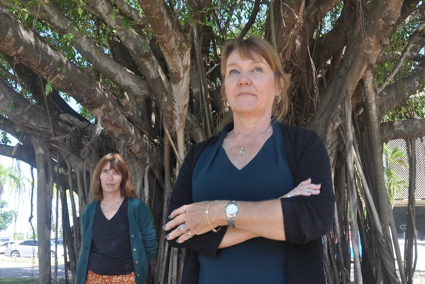 Two women stand under a shady tree.