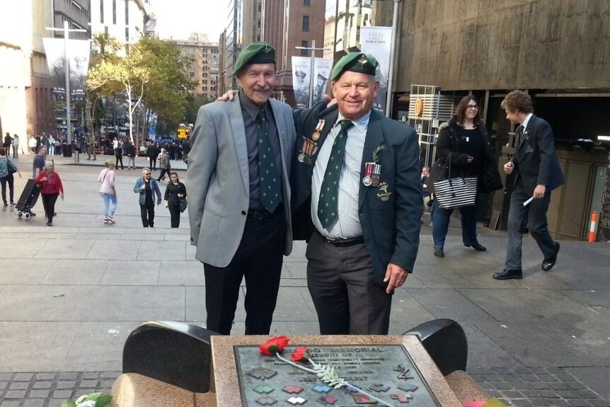 Two men aged in their 70s wear war medals and stand in front of memorial