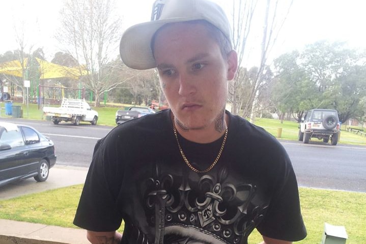 A young man with a white cap, tattoos and black t-shirt standing near a footpath
