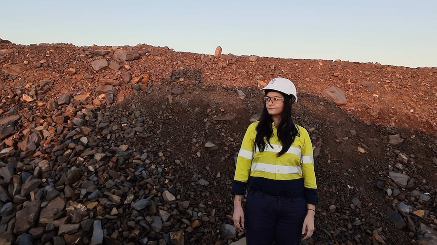 A woman in a yellow shirt, hard white safety hat and clear safety goggles stands in front of a pile of red dirt and rocks.
