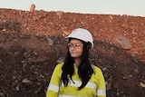 A woman in a yellow shirt, hard white safety hat and clear safety goggles stands in front of a pile of red dirt and rocks.
