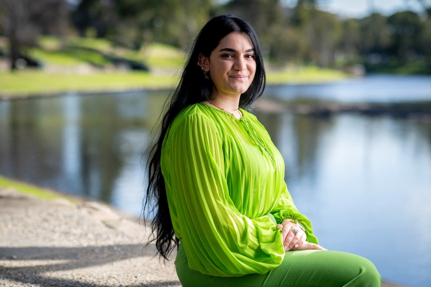 A young woman with long black hair smiles in a green dress in front of a lake