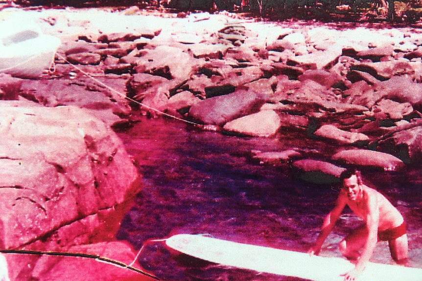 An older photo showing a rocky shore and man standing beside a surfboard, smiling.